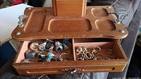 Open wooden jewellery box showing two-section compartment filled with assorted metal rings, some with colored stones.