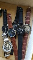 Six men's wristwatches laid side by side showing variety of bands, dial styles, and brand markings. Watches not tested. Shows black, brown leather straps, and metal bracelets.
