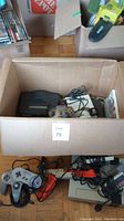 Cardboard box on floor containing black N64 console, cables, controllers, SNES cartridges visible inside, two SNES controllers and NES unit with Zapper on floor
