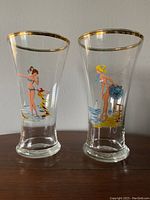 Pair of clear pilsner glasses with gold rims and mid century pin-up prints on the front, standing on a wooden surface.