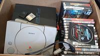 Cardboard box containing Sony PS2 fat console, white PSOne console, stack of game cases