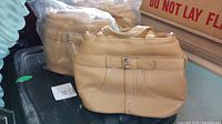 Three tan Gilbro handbags on a black surface with two still in clear plastic packaging and one unpackaged showing its front with a belt buckle detail.