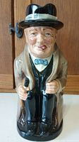 Front view of the Royal Doulton Winston Churchill ceramic character jug, showing detailed facial features, black hat with white band, beige coat, and blue bow tie.