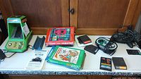 Wide view of the lot items on white table showing Sega Frogger handheld, Intellivision boxes and cartridges, manuals, Microsoft controller and handheld poker game.