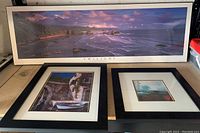 Three framed decorative pictures arranged side by side: a large panoramic coastal seascape, a smaller picture of a stone archway, and a framed sailboat picture.