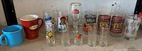 Overview of 12 assorted vintage drinking glasses and mugs on a shelf showing various brands and designs