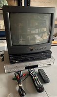 Photo of the 13-inch black Magnavox CRT TV with built-in speaker and stacked DVD VHS combo player below, showing connection cables and remotes in front.