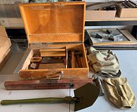 Wooden toolbox open showing tools inside, large wooden level, green military shovel, and two canvas military bags outside.