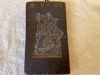 Rectangular brown wax sculpture/cookie mold with detailed relief of a woman at a spinning wheel, with inscription 18.1.P.E 04 visible on top.