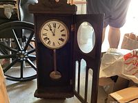 Full view of antique wooden pendulum wall clock with door open showing pendulum and clock face