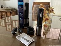 Photo showing Oster cordless electric wine opener, charging base, power adapter, user manual and original box on table.