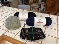 Photo showing 11 vintage crochet hooks in a black case, two full skeins of Big Twist yarn in navy blue and black, and one partial gray skein on a kitchen countertop.