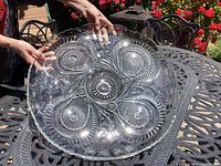 Photo showing entire round cut glass serving tray with intricate designs and scalloped edge, held by hands for scale on metal outdoor table.