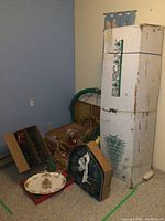 Large artificial Christmas tree in tall BarcaNia box with various holiday decorations including wreaths, garland, lights, and a ceramic tree on floor beside it.