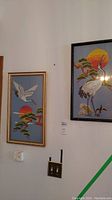 Two framed needlepoint crane art pieces hung on a wall, side by side, showing general view and size comparison.