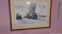 Framed print showing a steam train in a snowy mountain setting, with tan mat and wooden frame. Wall color is pink.