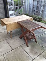 Wide shot of solid pine drop-leaf table next to wooden red outdoor cart on patio stone flooring with wooden fence background