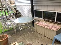 Full view of patio furniture including round table, stack of 3 metal chairs, and beige deck box with cushions inside.