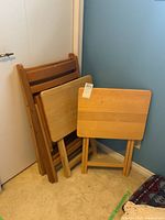 Two wooden folding TV tables and two wooden folding chairs folded and stacked against a wall