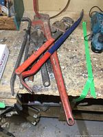 Vintage metal tools including red pipe wrenches, adjustable wrench, wrecking bars on wooden workbench.