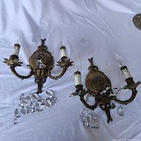 Pair of brass two-arm wall sconces with candle-shaped bulbs and loose hanging crystal prisms on white fabric background.