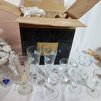 Photo showing multiple glasses including historic Federal Atomic Ameoba Boomerang glasses featuring blue amoeba and boomerang designs on clear glass, assorted shot and cocktail glasses, plus a vintage Libbey Gold Pine Cone frosted pilsner glass with gold pine cone decoration.