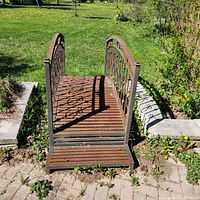 Front view showing the entire iron metal bridge with wooden slat walking surface and decorative railings with scrollwork, with some surface rust visible.