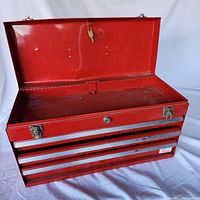Red metal toolbox with open lid showing empty top compartment and three closed drawers with aluminum handles. Key visible hanging from lid lock.
