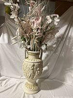 Full view of large Romanesque style cream-colored vase filled with artificial white and pink flowers and greenery, set against a white backdrop.