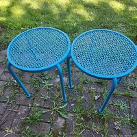 Two blue metal outdoor side tables on grass and stone ground, showing their circular lattice tops and legs.