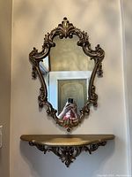 Gold ornate wall mirror with intricate baroque style frame hanging above matching semi-circular entryway shelf, both mounted against a wall.