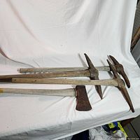Lot showing three pickaxes and one axe, all with wooden handles and rusted metal heads, laid out on a white sheet.