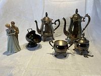 Full composition showing two silver-plated tea sets with teapots, creamers and sugar bowls, black sugar bowl with spoon, and porcelain figurine of a decorated couple holding miniature tea set pieces