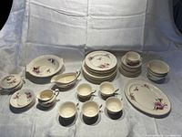 Full view of entire china set arranged on white cloth showing multiple cups, plates, and serving pieces with pink carnation floral decoration and gold trim edges.
