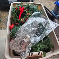 Overhead view of large beige plastic bin filled with green garlands, a small wreath decorated with red bows and berries, and a wrapped lighted garland.