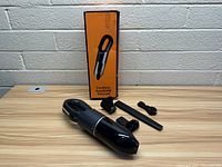 Cordless handheld vacuum with accessories and original box displayed on a wooden table against a white brick wall background.