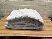 Folded white king size mattress topper placed on wooden table against beige brick wall, showing quilted pattern and thick cushioning.