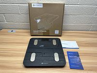 Smart fitness scale in front of original box with user manual and informational card on wooden table.