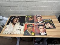 Top view of large Elvis poster and five Elvis Presley magazines laid out on a wooden table against a grey brick wall.