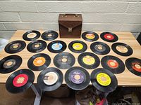 Sixteen 45 RPM records arranged in front of a brown carrying case on a wood table, showing varied label colors and designs