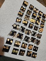 Photo of a large assortment of earrings arranged on a grey surface showing multiple pairs of gold-toned earrings with various designs and mounted on black Madeline branded cards.