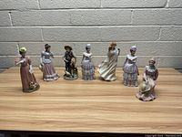 Seven porcelain figurines arranged in a row on a wooden surface against a white brick wall, showing full front view of each.