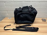 Front and side view of black multi compartment tote carrying bag with multiple zippered compartments and detached shoulder strap laid on wooden table against brick wall.