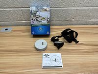 Top-down photo of the transmitter device, collar strap, instruction manual, and product box on a wooden surface.