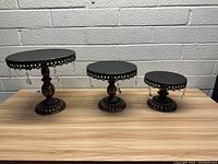 Set of 3 black metal pedestal stands on wooden surface against gray brick wall, arranged by size from largest to smallest
