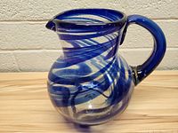 Front angle of the clear glass pitcher with blue swirls showing bulbous body, curved handle, and pouring spout placed on wooden surface against brick wall background.