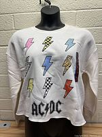 Front view of white womens sweater with AC/DC logo and colorful lightning bolts, displayed on mannequin against a cinder block wall.