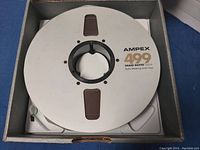 Photo of a silver metal reel with Ampex 499 Grand Master Gold branding and text, housed inside a gray cardboard box insert.