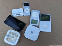 Photo showing untested Apple devices including an iPhone 4 black with rear glass, three iPods (Classic and Nano), a 30-pin dock adapter, a white square Apple device with logo, and Apple earbuds in a plastic case.