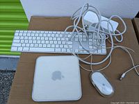 Photo showing Apple Mac Mini, wired keyboard, mouse, and cables on brown surface.